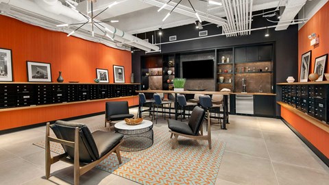 Looking in to the Clubroom at Wrigleyville Lofts. There are club chairs in a circle ahead with a high-top table behind. The far wall has a mounted television with beer taps beside. The side walls are orange with mailboxes built-in for residents.