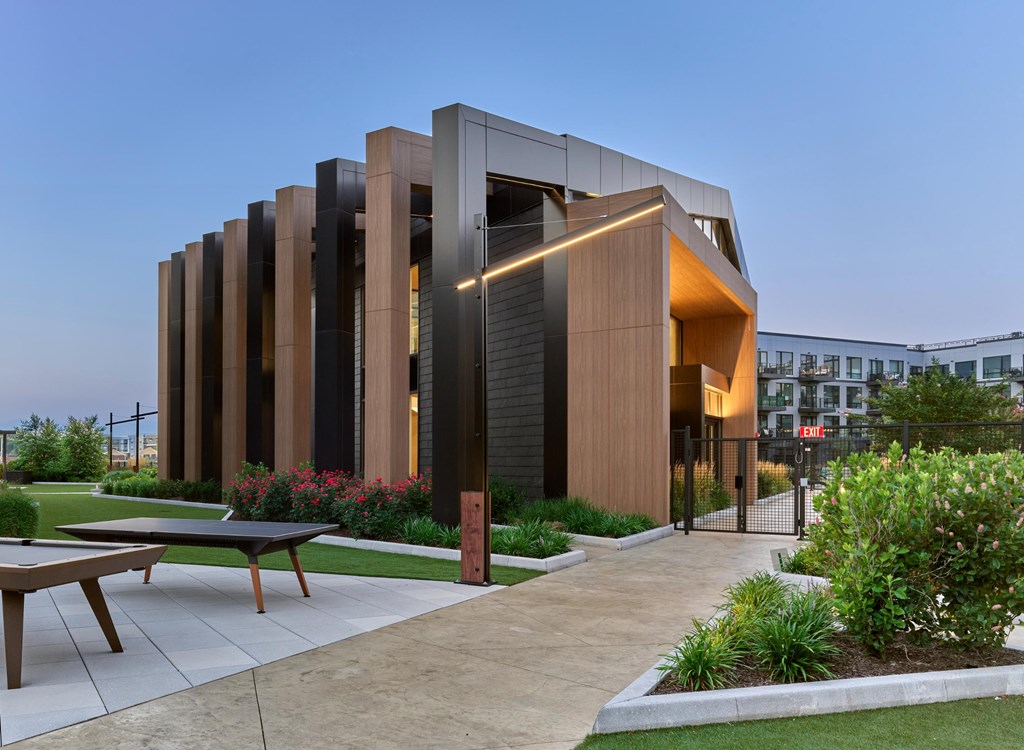 A modern building with a glass facade and a wooden exterior is surrounded by a garden with a picnic table.