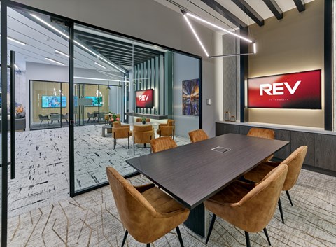 A conference room with a long table and chairs with a glass wall and a REV sign on the wall.