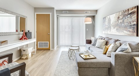 A bright living room with a small sectional, chair. A sliding glass door with blinds