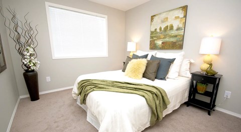 A bedroom with queen sized bed and two nightstands. a large window with blinds on one wall