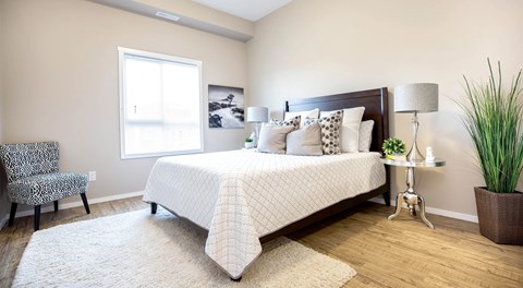 A bedroom with queen sized bed, night stands and a chair. A large window with blinds on far wall.