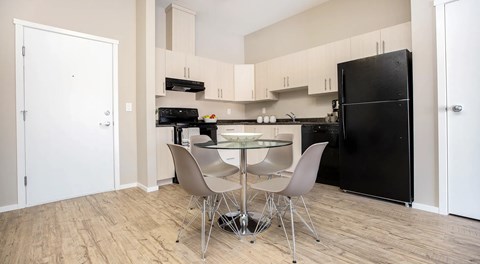 An open kitchen, with light cabinets, dark counters and black appliances. A round table with four chairs around it sits in the centre