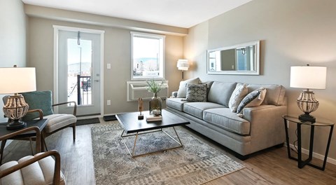 A bright open living room with a couch, chairs, coffee table and end tables. Against outer wall a glass patio door, a window with AC unit below