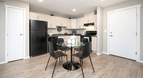 An open kitchen with light cabinetry and black appliances, a small pantry next to the fridge and a table for four in the centre