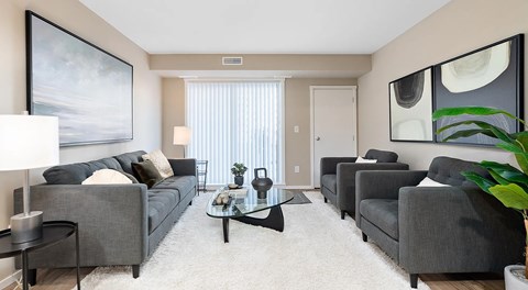 An open living room with a grey couch on one side and two arm chairs on the other, separated by a coffee table in the middle. A glass sliding door leading to a balcony