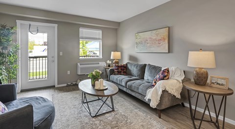 An open living room with a couch, large chair, coffee table and end tables. A patio door with glass insert next to a window and wall mounted AC unit