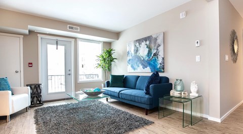 A bright open living room with a couch, side table, coffee table and chair sit next to a large window and blacony door with window and blinds