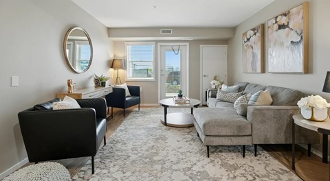 A living room with a couch and some chairs, a coffee table sits in the centre with a window and patio door behind