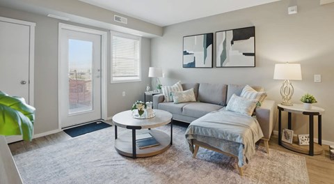 A living room with a small grey sectional, side and coffee table with a window and balcony door