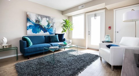 A bright open living room with couch, coffee table and chair. A bright window sits next to a glass balcony door