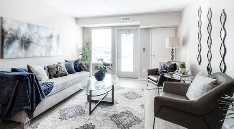 A living room with a couch, a coffee table, and two chairs. A large window and balcony door sit on the exterior wall