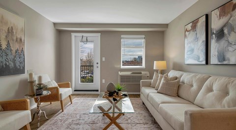 A bright open living room with a couch, coffee table and chairs, balcony door, window and AC unit