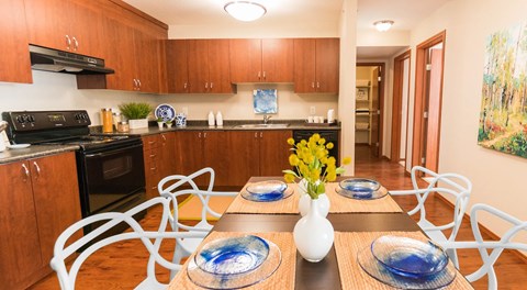 A kitchen with warm wood cabinets, dark counters and black appliances, a long rectangular table runs down the centre of the kitchen