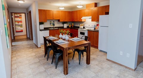 A warm toned kitchen with wooden cabinets, dark counters and white appliances. A large rectangular table sits in the centre.