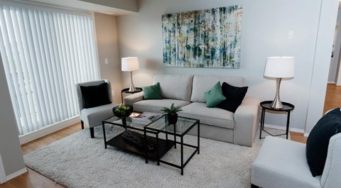 A bright open living room with a couch, chairs and coffee tables, two end tables and a large balcony glass sliding door with blinds