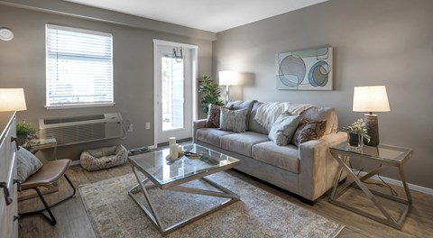 An open living room with a couch, coffee table, chair and end tables. One wall has a balcony door, medium sized window and an AC unit below