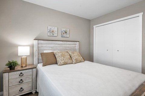 A bedroom with a white bed, a wooden nightstand with a plant and a lamp, and two framed pictures on the wall.