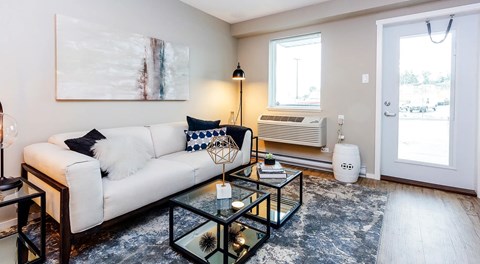 A living room with couch, end tables and coffee table. A wall with a medium sized window with AC unit below and a patio glass door