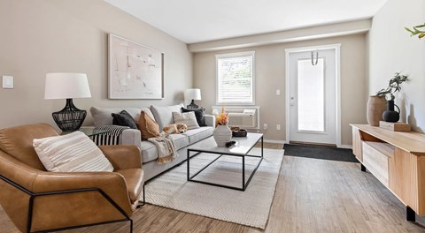A bright open living room with a couch, chair, coffee table and tv stand. A glass balcony door, with window and AC unit next to it
