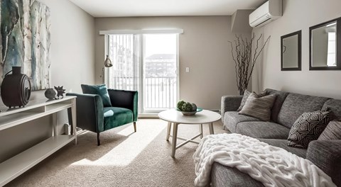 A bright living room with couch, coffee table, chair, and glass sliding door with blinds and wall mounted AC unit