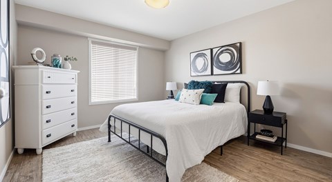 A bedroom with a queen sized bed, a white dresser, night stands and large window