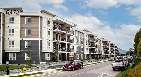 An apartment building with lots of windows and balconies sits next to a street with street parking