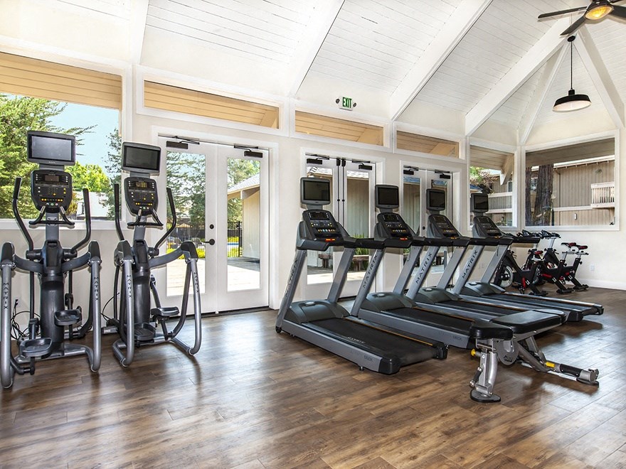Three Bedroom Apartments in Santa Cruz CA - Cypress Point - Gym with Black Treadmill and Elliptical Machines with Screens, Wood Floors, and Windows Under a Vaulted Ceiling with Lighting and Fan