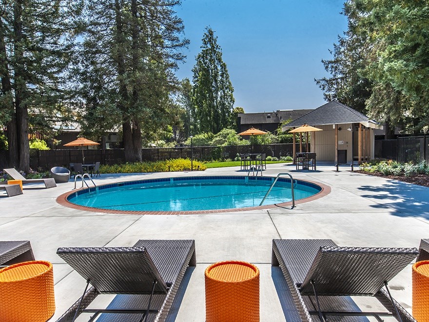 Circular pool surrounded by deck chairs, orange tables, trees and grass.