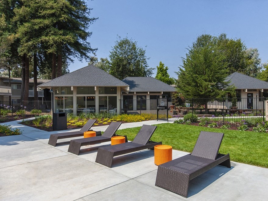 Concrete pool deck with brown deck chairs and orange tables. Grass, club house and lounge behind them.