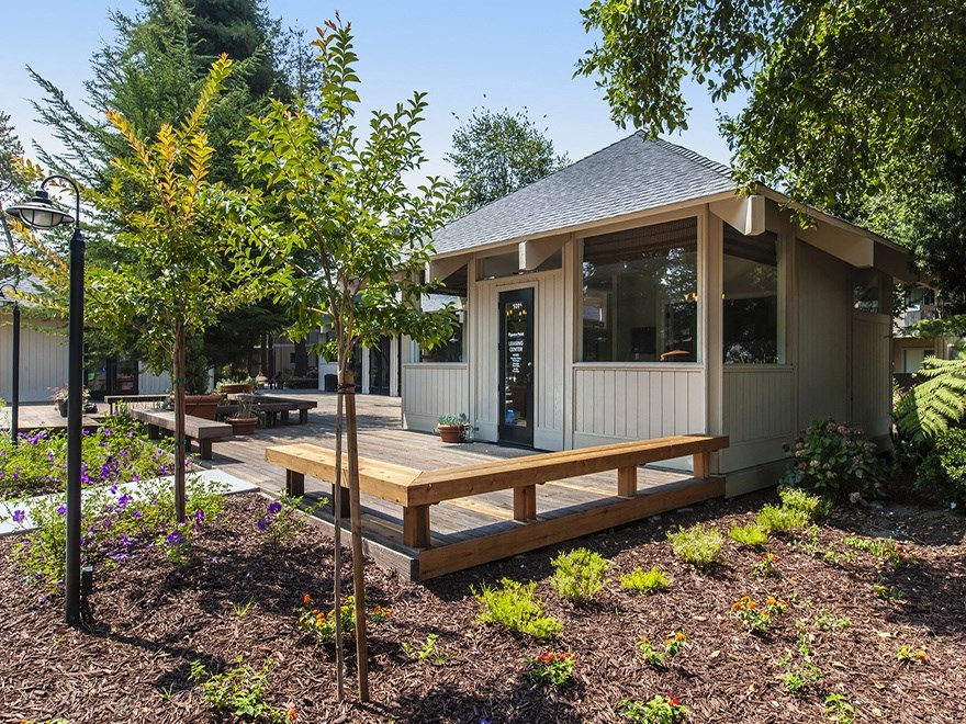 Trees and flowers leading up to lounge building and patio with wooden benches as outdoor seating attached to lounge.