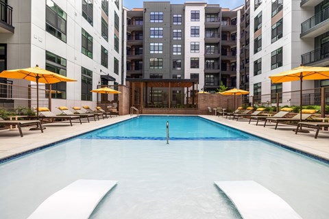 A swimming pool surrounded by lounge chairs and umbrellas in front of apartment buildings.