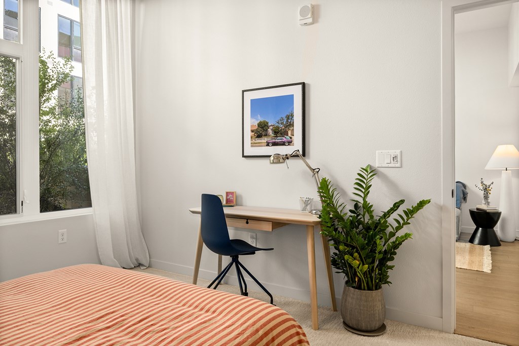 A bedroom with a bed, desk, chair, and potted plant.