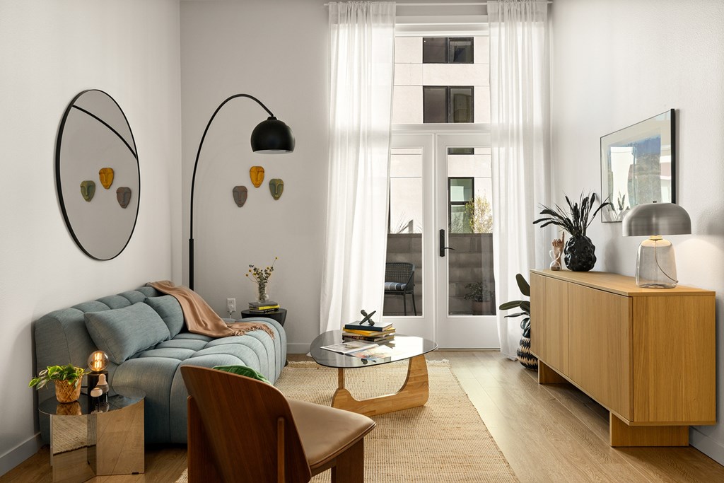 A living room with a blue couch, a wooden chair, a round table, and a large window with white curtains.