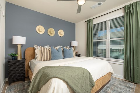 Calm modern bedroom with a white bed, sage-green throw and curtains, blue accent wall, moon wall art, bedside lamps, and wide windows.