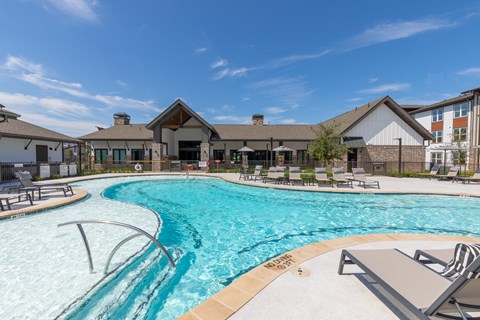 A bright resort courtyard features a turquoise swimming pool, curved shallow entry, lounge chairs, and lodge-style buildings under a blue sky, creating a relaxed, welcoming mood.