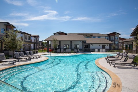 A bright apartment complex surrounds a turquoise swimming pool, with lounge chairs lining the curved deck beneath a clear blue sky, creating a relaxed, inviting mood.