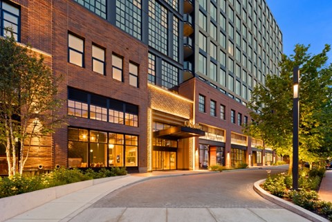A modern building with a brick facade and large windows is illuminated from within, with a pathway leading up to the entrance.