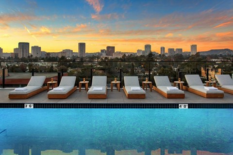A pool with lounge chairs and a city skyline in the background at sunset.