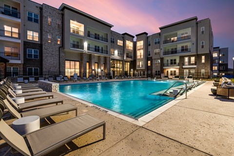 A swimming pool surrounded by sun loungers in front of apartment buildings.