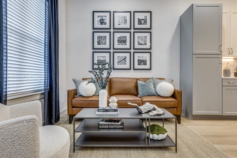 A living room with a brown couch, a glass coffee table, and a wall with framed pictures.