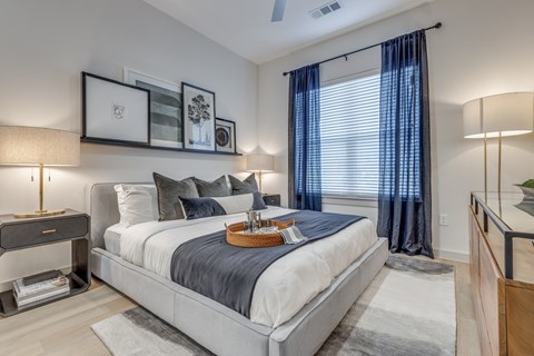 A bedroom with a large bed, a nightstand with a lamp, and a window with blue curtains.