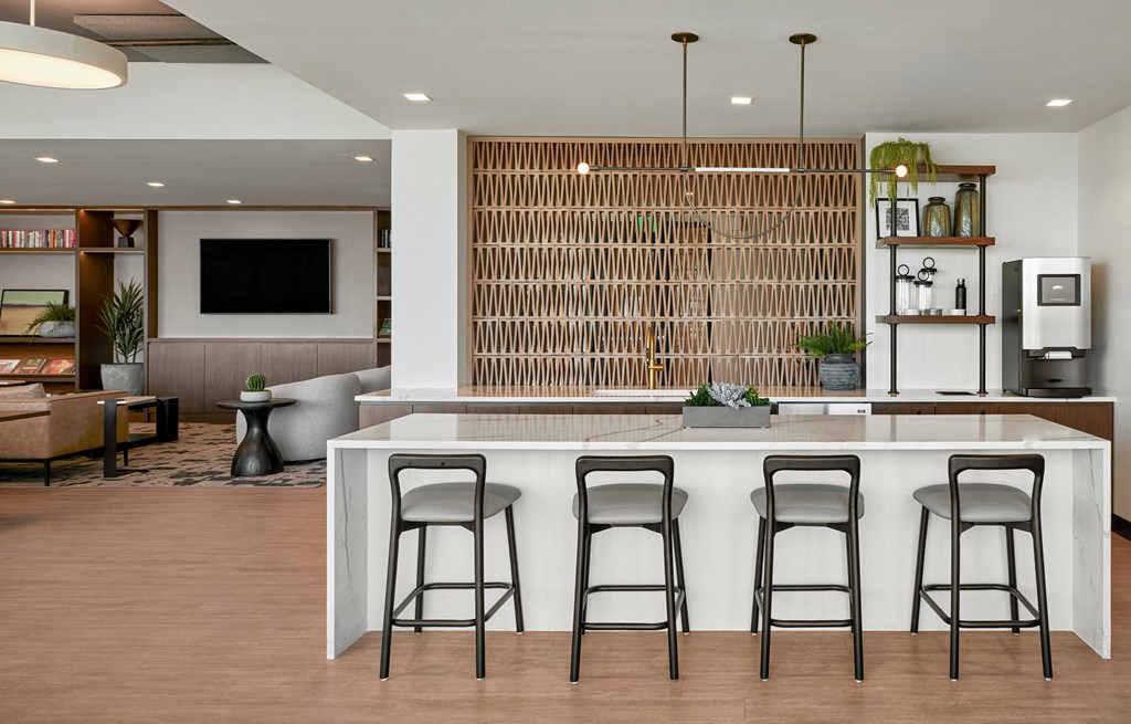 an indoor kitchen with a counter and three stools, coffee machine, ceiling lighting