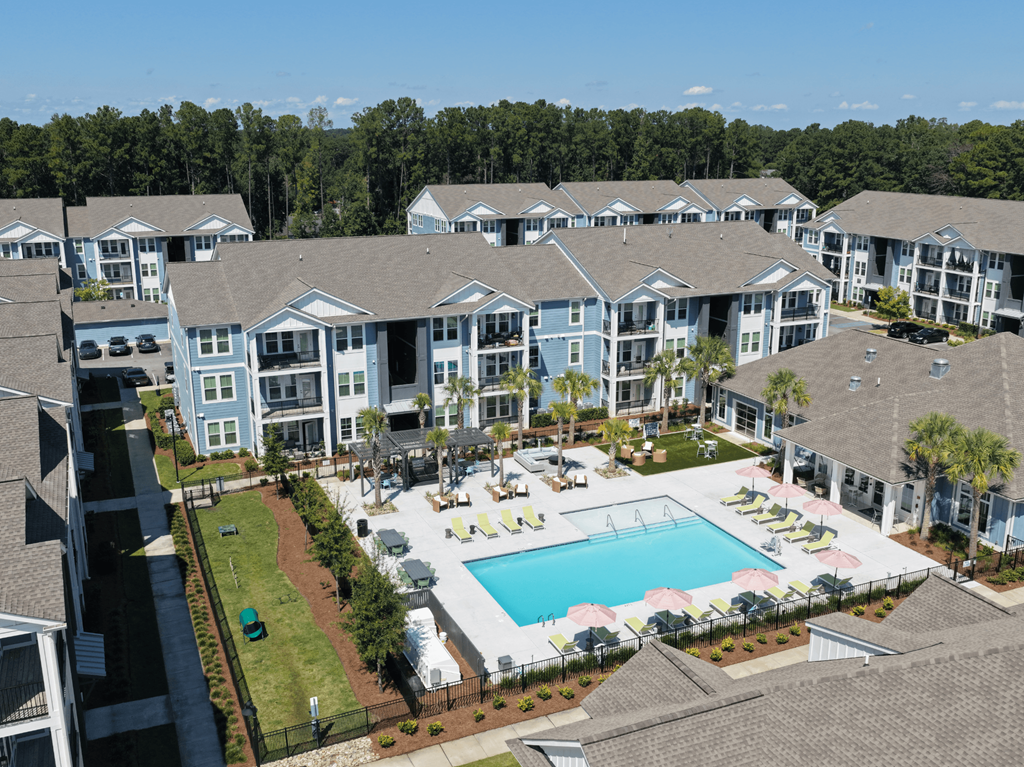 A large swimming pool surrounded by lounge chairs and umbrellas is the centerpiece of a resort-style apartment complex.