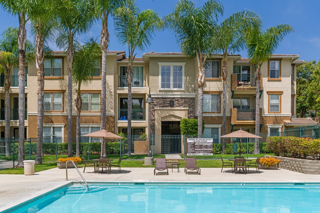 A swimming pool is surrounded by lounge chairs and umbrellas in front of a large apartment building.