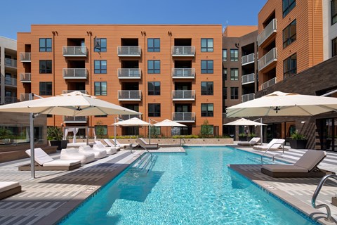 A large swimming pool surrounded by lounge chairs and umbrellas in front of a multi-story apartment building.