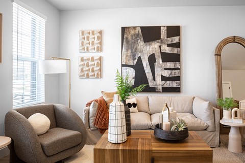 A living room with a grey armchair, a wooden coffee table, a large abstract painting on the wall, and a mirror on the right side.