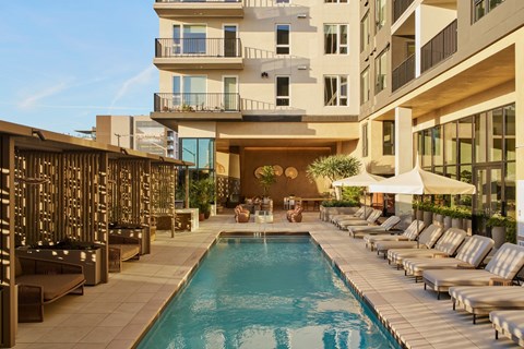 A large swimming pool surrounded by lounge chairs and umbrellas in front of a modern building.