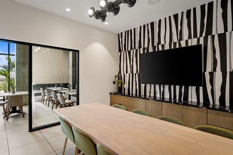A conference room with a long table and chairs, a television on the wall, and a glass wall leading to an outdoor dining area.