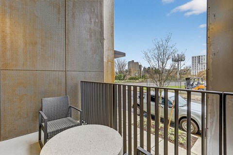 A chair and table are on a balcony with a view of a parking lot and buildings.